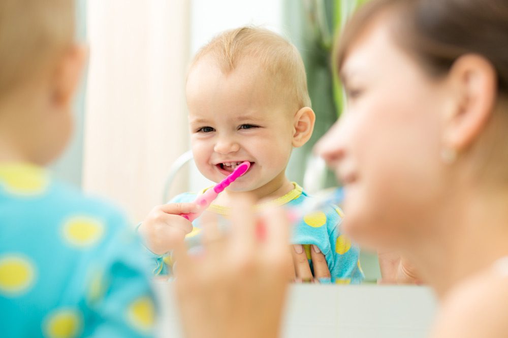 A toddler who is a patient at West Richland Family Dental Care brushes her teeth with her mother’s help.